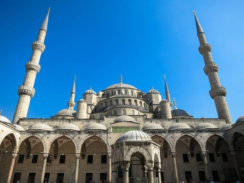 Je contemple la Mosquée bleue depuis sa cour intérieure à Istanbul, une mosquée ottomane du XVIIe siècle avec six minarets, un dôme central et une fontaine d'ablutions sous un ciel bleu éclatant.