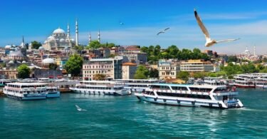 I am on a boat in Istanbul, facing ferries sailing on the Golden Horn, with the Süleymaniye Mosque, a 16th-century Ottoman mosque, dominating the hill in the background, and seagulls flying in the blue sky.