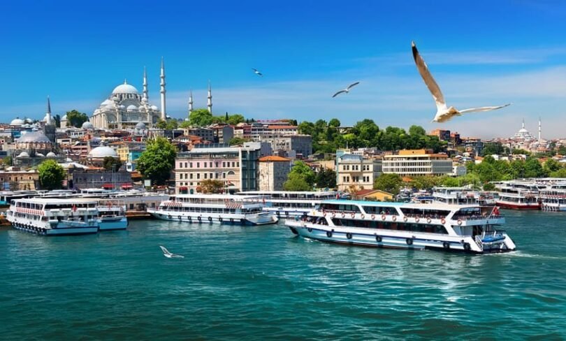 I am on a boat in Istanbul, facing ferries sailing on the Golden Horn, with the Süleymaniye Mosque, a 16th-century Ottoman mosque, dominating the hill in the background, and seagulls flying in the blue sky.