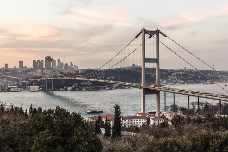 Je contemple depuis une colline le pont du Bosphore à Istanbul, un grand pont suspendu reliant l'Europe et l'Asie, avec un ferry qui passe en dessous et des gratte-ciel en arrière-plan sous un ciel nuageux.