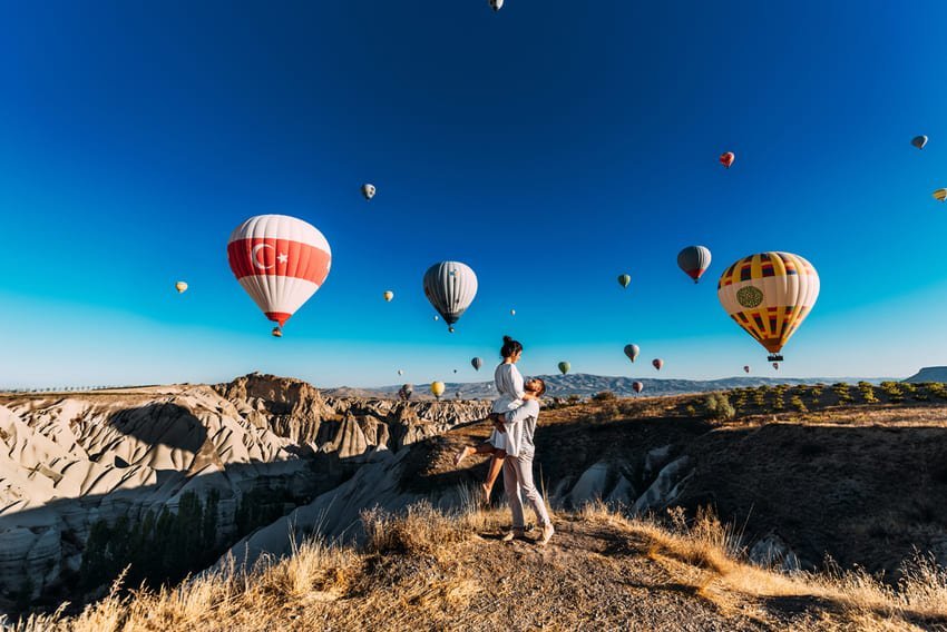 Un voyageur vêtu de vêtements légers se tient debout sur un terrain herbeux, face à 12 montgolfières colorées flottant au-dessus des cheminées de fées beiges de la Cappadoce. Le ciel bleu clair et les formations rocheuses creuses s'étendant à l'horizon symbolisent la rencontre entre l'héritage ancestral et les loisirs aériens modernes.