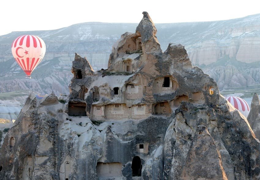Forteresse troglodyte couronnée de deux montgolfières roses flottant au coucher du soleil. Le château transparent domine la vallée de Cappadoce avec ses formations géologiques calcaires sculptées, ses tunnels intérieurs et ses grottes habitées.