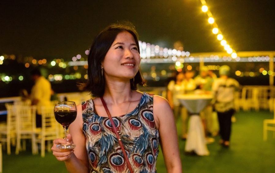 Femme tenant un verre de vin sur le pont d'un bateau, éclairage nocturne, guirlandes lumineuses, invités en arrière-plan et vue sur la ville ou le pont du Bosphore.