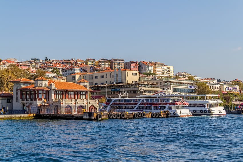 Il molo di Eminönü visto dalla barca, edifici coloniali ottomani multicolori di 3-4 piani che si affacciano sulla terrazza del ristorante. Il ponte di Galata in metallo che collega Europa e Asia sullo sfondo, traghetti turistici colorati, negozi del porto. Centro storico bizantino del I secolo, nodo marittimo transcontinentale, moderno snodo ferroviario e tranviario dove cucine galleggianti e taxi-traghetti animano l'orizzonte urbano.