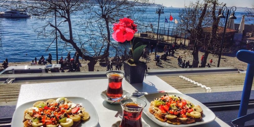 Une table sur la terrasse avec deux assiettes de gaufres garnies de fruits et deux verres de thé, avec en toile de fond le Bosphore animé. Je suis assis en face d'un café avec une vue panoramique sur les berges et les bateaux qui passent.