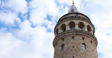I am facing the Galata Tower seen from a low angle, with its arches and spire; built in 1348, it peaks near 67 m, a historical lookout of Istanbul.