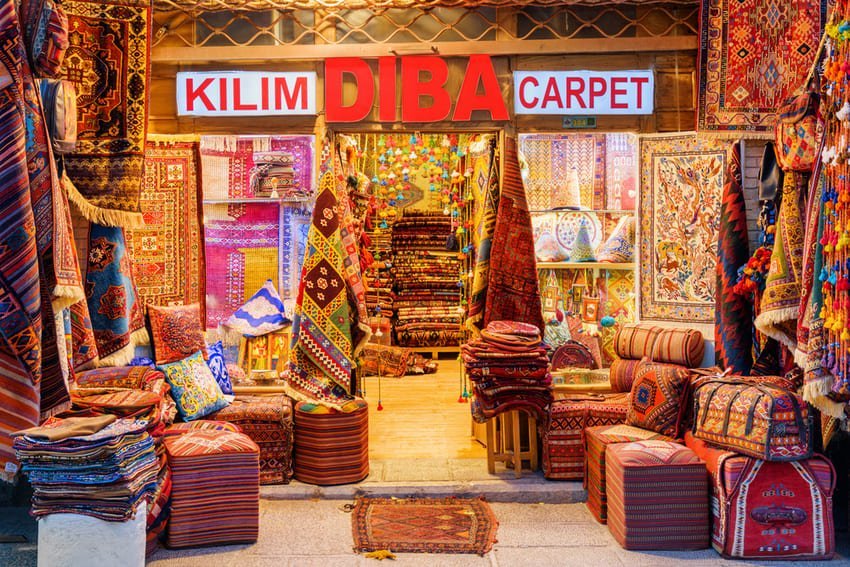 Front of a carpet shop in Istanbul with stacks of colourful kilims and a neon sign above the entrance. I am looking at traditional flatweaves from Anatolia, prized for their geometric patterns and natural dyes.