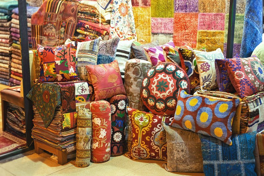 Shop window in Istanbul displaying stacks of decorative cushions with floral and geometric embroidery in bright colours. I am looking at pieces inspired by suzanis and kilims, some made from reassembled antique carpets.