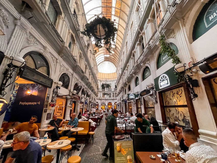 Passerella coperta decorata con tavoli allegri, volta in vetro e arredamento raffinato. Interni in pietra bianca, atmosfera calda tipica delle gallerie di Istanbul.