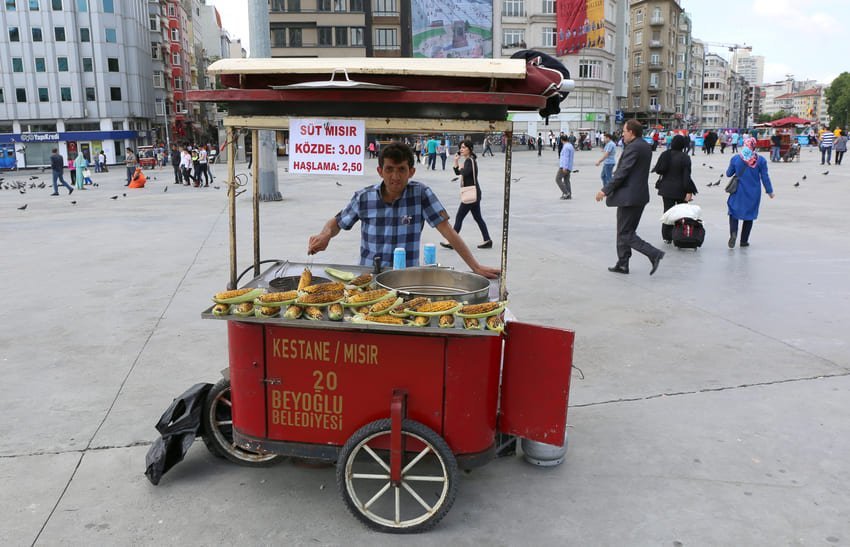 Venditore ambulante con un carretto rosso in una grande piazza cittadina, che offre mais arrostito e bollito, con gente che si muove intorno a lui. Atmosfera vivace e tipica della città.