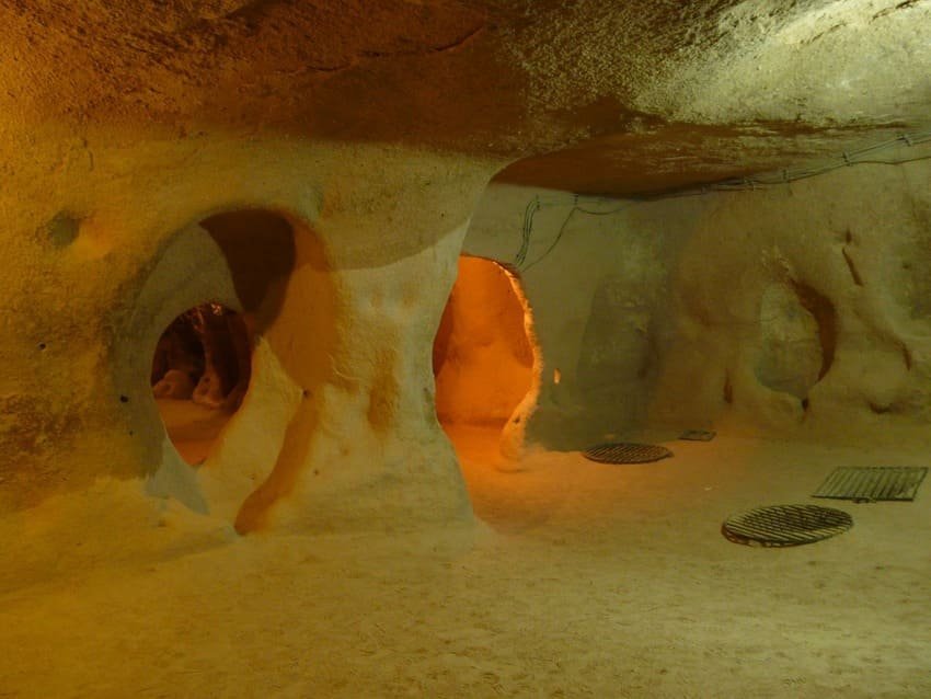 Intérieur d'un hôtel troglodyte avec des arches et des murs en tuf beige-gris, des fenêtres basses orange qui confèrent à l'habitat une ambiance lumineuse. Passage vers d'autres pièces souterraines, grilles d'aération au plafond. Hébergement historique millénaire converti en tourisme haut de gamme.