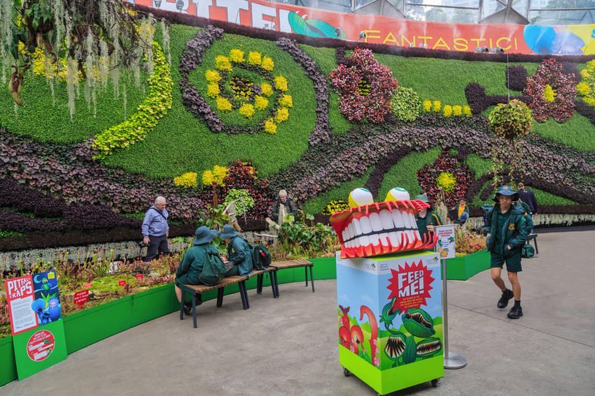 Mur végétal géant décoré de spirales de fleurs, de personnages assis et d'une mascotte colorée. Ambiance ludique et colorée de jardin intérieur.