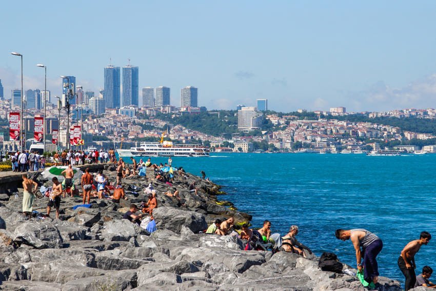 Spiaggia rocciosa di Kuzguncuk, bagnanti asiatici che si godono le acque turchesi del Bosforo di fronte ai grattacieli di Beyoğlu. Spiagge storiche in pietra, alberi costieri, negozi turistici tradizionali sullo sfondo. Quartiere costiero multiculturale di Üsküdar che conserva l'atmosfera dei villaggi ottomani con vecchie case in legno, caffè, moschee e chiese.