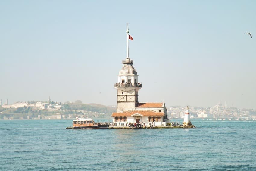 Je contemple la Tour de la Vierge, haute de 23 mètres, au crépuscule, avec une mouette qui vole devant elle, un ferry amarré et la silhouette de la Mosquée bleue et de Sainte-Sophie visibles à 2 kilomètres de là, sur la péninsule historique, sous un ciel doré.