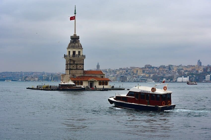 Je me trouve face à un bateau traditionnel naviguant vers la Tour de la Vierge à Istanbul, haute de 23 mètres et située à 200 mètres de la rive d'Üsküdar, avec la silhouette de la ville historique visible en arrière-plan sous un ciel nuageux.