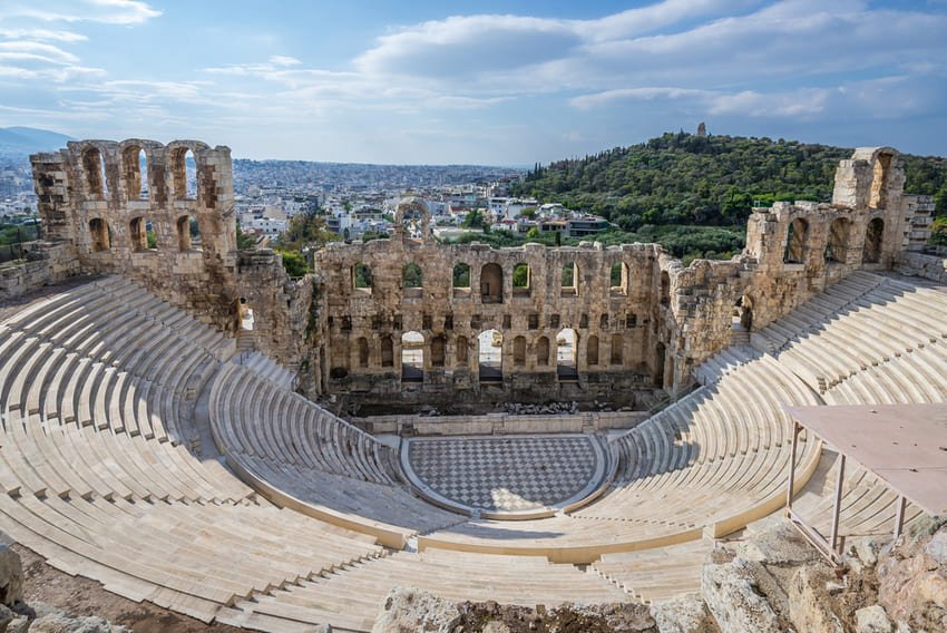 Je contemple l'Odéon d'Éphèse, un petit théâtre semi-circulaire construit entre 140 et 150 après J.-C., avec ses 22 rangées de sièges en marbre blanc restaurés en arc, sa scène à deux étages avec des colonnes corinthiennes rouges, son podium surélevé d'un mètre, sa capacité de 1 500 spectateurs, flanqué de hauts murs de pierre et de montagnes boisées en arrière-plan.