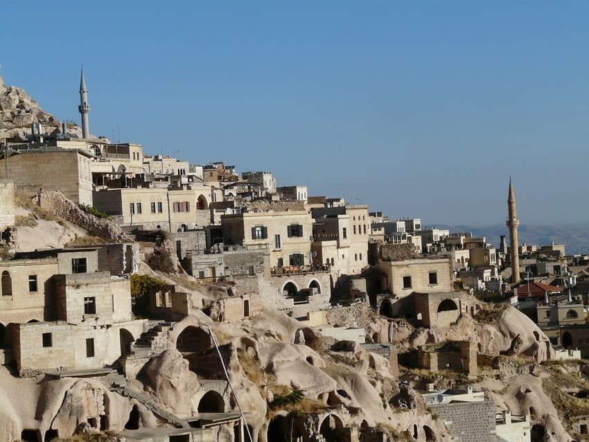 Le village d'Ortahisar, avec ses maisons en pierre beige et son château fort en tuf dominant. Maisons troglodytes, minarets en arrière-plan, paysage semi-aride de la Cappadoce s'étendant à l'horizon. Habitat millénaire perché, vue panoramique depuis le promontoire rocheux créé par l'érosion volcanique.