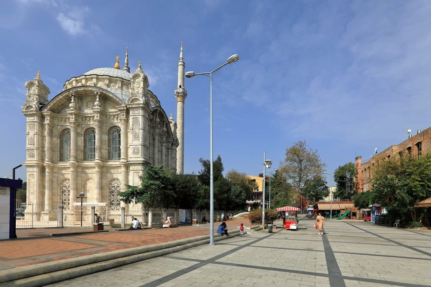 La moschea Mecidiye in marmo bianco sulla piazza pedonale di Ortaköy, a pianta quadrata in stile neobarocco, con due minareti slanciati e facciata adornata da sculture. Edificio del 1854-1856 progettato dagli architetti Balyan, domina un vivace quartiere commerciale con negozi, ristoranti e passanti. Fusione di stili ottomani ed europei che riflette la modernità del Tanzimat.