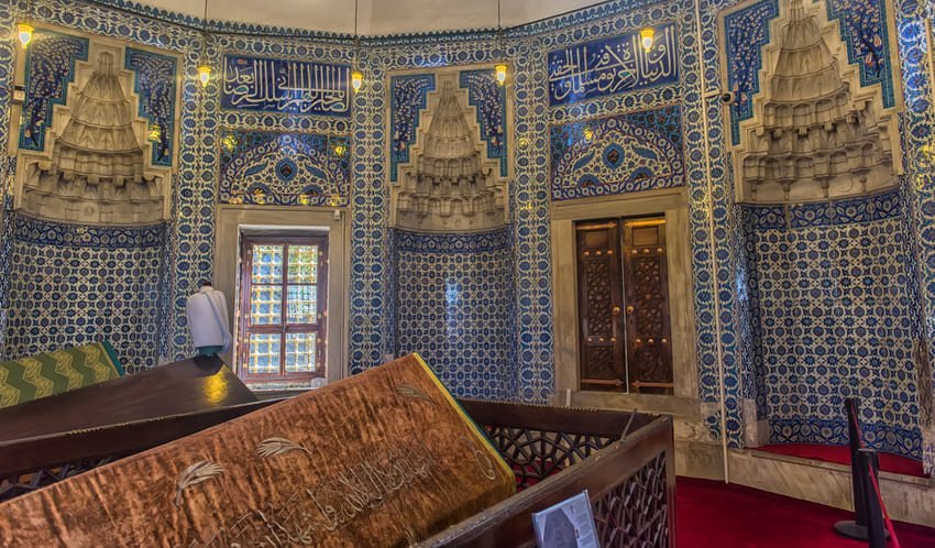 Je me trouve dans un mausolée ottoman décoré de carreaux d'Iznik du XVIe siècle, avec des murs entièrement recouverts de céramiques florales bleues et blanches, trois lustres pyramidaux dorés et des calligraphies arabes encadrant les portes et les fenêtres.