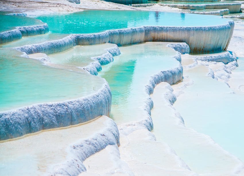Je contemple les bassins en cascade de Pamukkale, avec leurs eaux turquoise et bleues contenues par des bordures stratifiées de travertin blanc, formées par le dépôt de 499,9 milligrammes de carbonate de calcium par litre d'eau thermale entre 35 °C et 100 °C refroidissant à la surface.