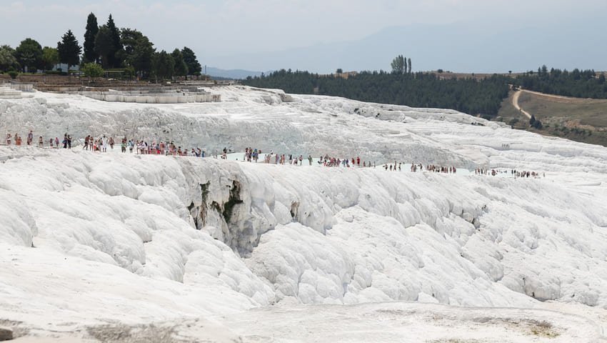 I discover the panorama of the white travertine terraces of Pamukkale extending over 2.7 kilometers long and 600 meters wide, with hundreds of visitors walking on the limestone formations peaking at 200 meters high and the Roman ruins of Hierapolis visible at the top.