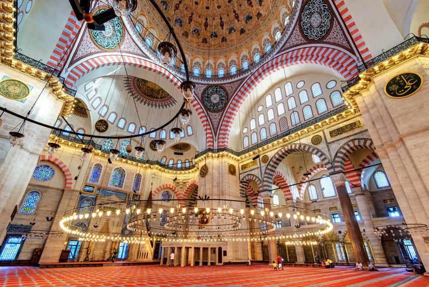 Je me trouve face à l'intérieur de la mosquée Süleymaniye à Istanbul, avec son imposant dôme central de 26,5 mètres de diamètre flanqué de demi-dômes, ses arches rouges et blanches caractéristiques et son immense lustre circulaire suspendu au-dessus du tapis de prière rouge.
