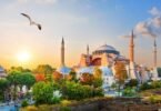 Visiting Istanbul. Panoramic view of the Hagia Sophia mosque in Istanbul at sunset, with turquoise domes, minarets, orange-leaved trees, seagulls and crowds in the foreground.