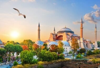 Visiting Istanbul. Panoramic view of the Hagia Sophia mosque in Istanbul at sunset, with turquoise domes, minarets, orange-leaved trees, seagulls and crowds in the foreground.