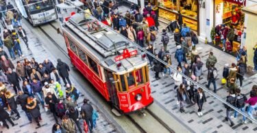 Historic red tram on Istiklal Avenue, crossing a dense crowd of passersby. Shops, cobblestones, and the lively atmosphere of central Istanbul.