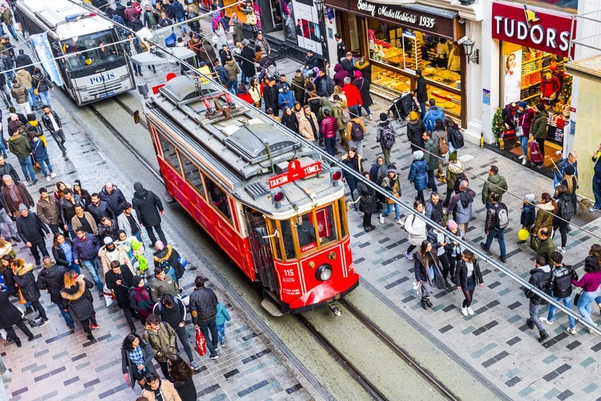 Tramway rouge historique sur l'avenue Istiklal, traversant une foule dense de passants. Boutiques, pavés et atmosphère animée du centre d'Istanbul.