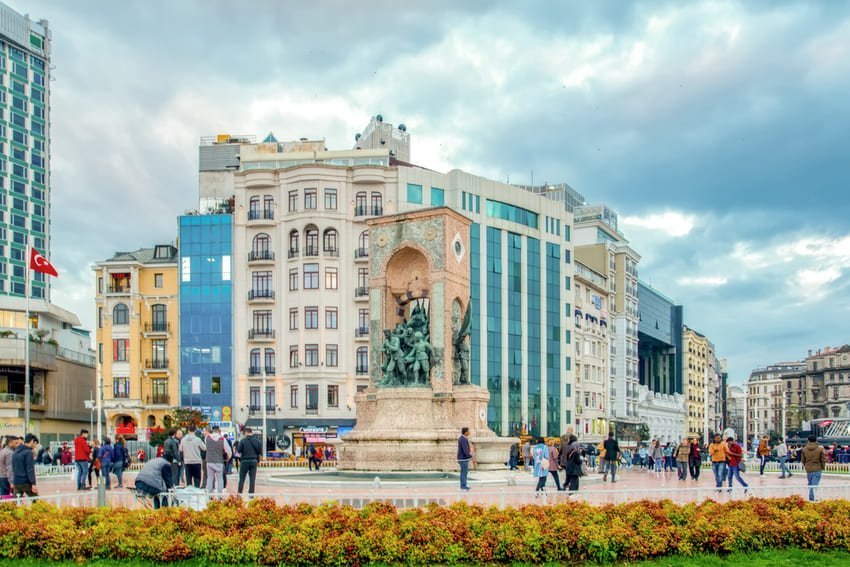 Monument de la République au centre de la place Taksim, encadré par les passants et les bâtiments modernes. Esplanade urbaine emblématique sous un ciel nuageux.