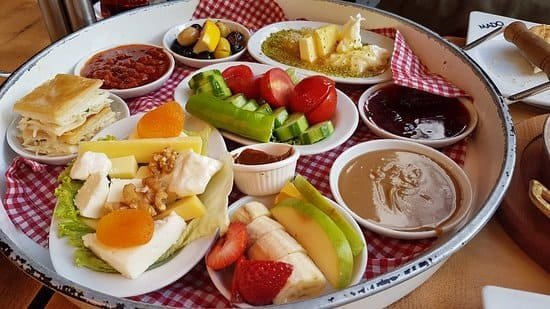 Je prends un petit-déjeuner turc à Istanbul, composé d'un plateau avec des fromages, du börek, des œufs, des olives, du miel, de la confiture, des concombres, des tomates et des fruits frais, le tout disposé sur des assiettes blanches à carreaux rouges.